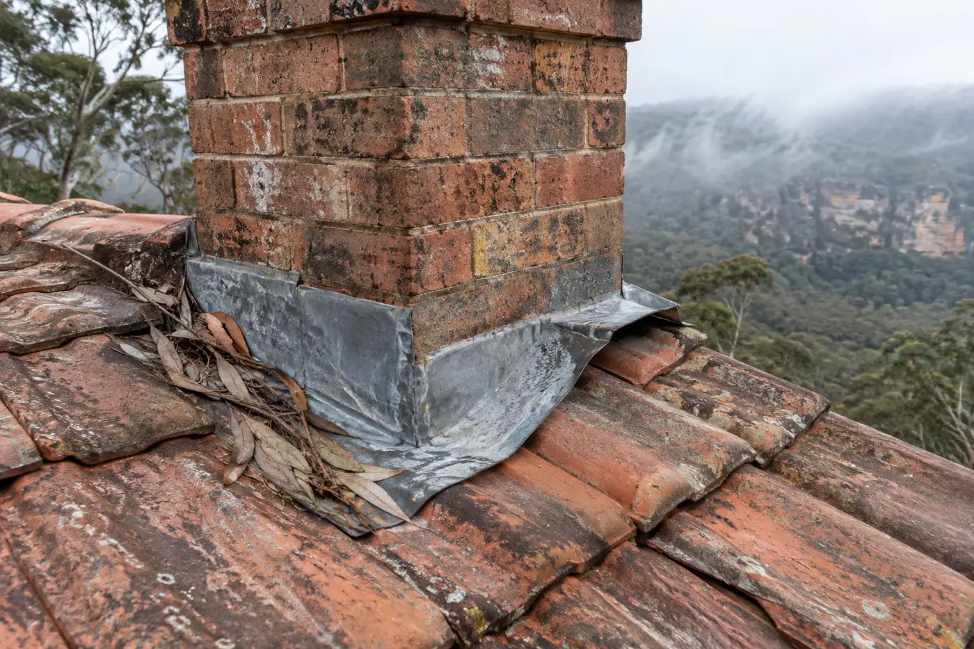 Lifted lead apron flashing at the base of a brick chimney on a weathered tile roof, with gum leaves caught on the uphill side